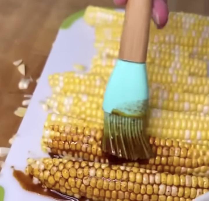 Ears of corn on the cob being brushed with a dark sauce on a cutting board