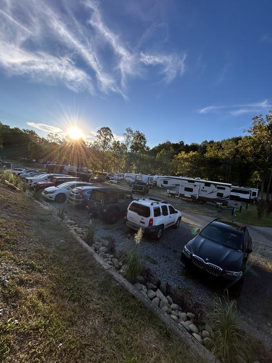 The sun breaking over the trees above a row of parked RVs and cars